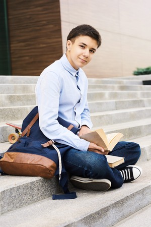 Portrait of smart teenage boy sitting on steps of school building looking at camera while reading books for studyingの写真素材