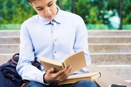 Portrait of smart teenage boy reading books for studying outdoors on stone steps of school buildingの写真素材