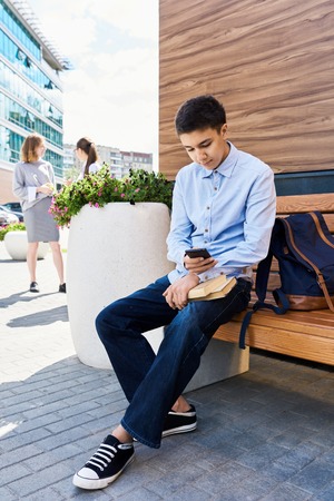 Portrait of teenage schoolboy relaxing on bench outdoors using smartphone and reading messagesの写真素材