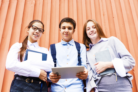 Portrait of three smiling schoolchildren holding books looking at camera standing against bright orange wallの写真素材