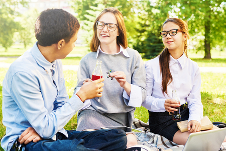 Group of smiling schoolchildren enjoying picnic on green lawn, chatting during breakの写真素材