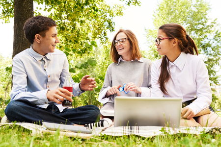 Group of modern schoolchildren enjoying picnic on green lawn, chatting and using laptopの写真素材