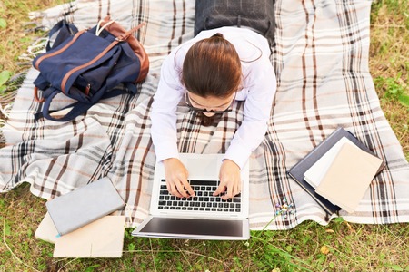 Above view of schoolgirl using laptop lying on blanket outdoors and studying during breakの写真素材
