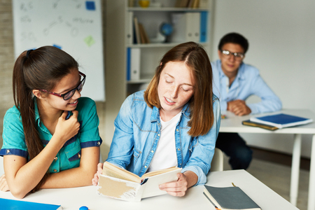 Portrait of two teenage girls reading book at desk together during lesson in modern classroomの写真素材