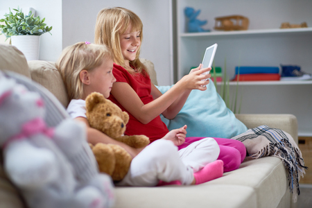 Profile view of joyful little girl showing her sister funny photo on digital tablet while they spending free time together at homeの写真素材
