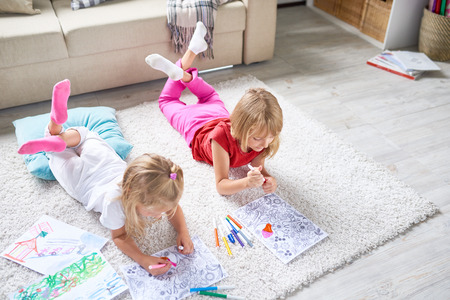 High angle view of adorable little sisters focused on painting coloring book with felt-tip pens while lying on floor in modern living roomの写真素材