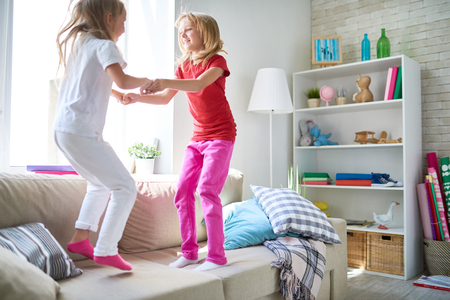 Two fidgety children holding hands and jumping on cozy couch while enjoying each others company in spacious living roomの写真素材