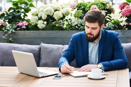 Portrait of contemporary bearded businessman working at table in comfortable cafe lounge outdoorsの写真素材
