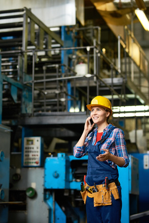 Portrait of young woman working in modern factory speaking by phone between rows of machines and smilingの写真素材