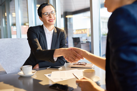 Waist-up portrait of beautiful middle-aged entrepreneur shaking hands with her business partner as sign of successful negotiations, interior of spacious cafe on backgroundの写真素材