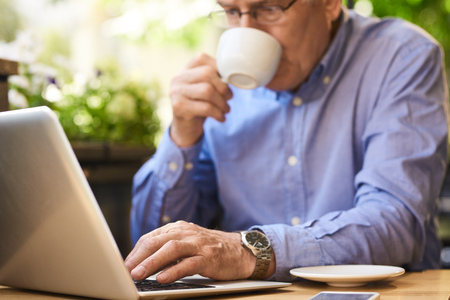 Closeup image of senior man using laptop computer enjoying coffee break in outdoor cafe loungeの写真素材