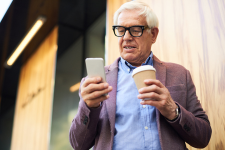 Portrait of modern senior man using smartphone outdoors, in city street holding coffee cup in one handの写真素材