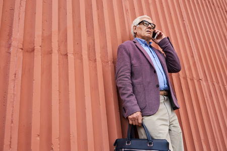 Low angle portrait of trendy senior man speaking by smartphone standing by bright orange wall outdoorsの写真素材