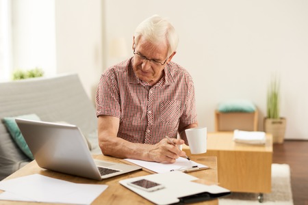 Portrait of senior man working with laptop at home counting finances and drinking coffeeの写真素材