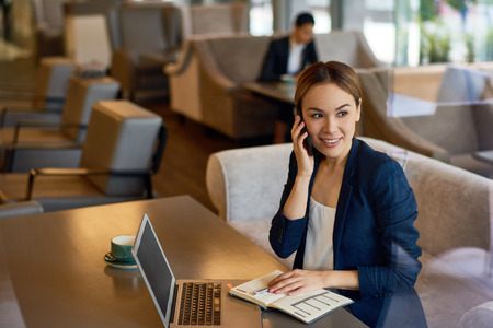 Attractive young manager talking to her business partner on smartphone while sitting at cozy small coffeehouse, waist-up portraitの写真素材