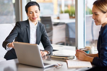 Attractive middle-aged businesswoman sharing ideas with her Asian colleague while gathered together in cozy restaurant with panoramic windowsの写真素材