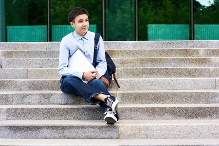 Full length portrait of handsome teenage student looking away with warm smile while sitting on stairs of modern school building, he holding laptop in handsの写真素材