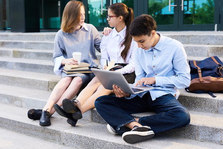 Teenage students having fun at break: pretty girls chatting animatedly with each other while sitting on stairs of modern school building, their handsome classmate browsing Internet on laptopの写真素材