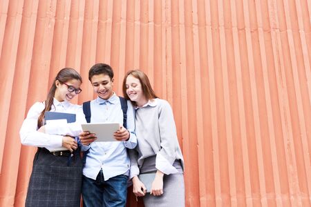 Low angle view of joyful teenage friends having fun outdoors: they watching funny video on digital tablet while standing at red concrete wallの写真素材