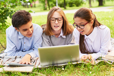 Enthusiastic group of teenage students gathered together in sunny green park and using laptop in order to prepare for exams, blurred backgroundの写真素材
