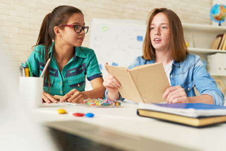 Pensive fair-haired student expressing her point of view concerning topical question while having class, her female classmate listening to her with interestの写真素材