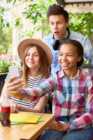Multi-ethnic group of three  happy students taking smartphone selfie in outdoor cafe on sunny summer dayの写真素材