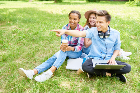 Portrait of three happy young people sitting on green lawn with laptop chatting and smiling enjoying break on summer dayの写真素材