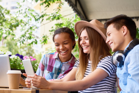 Multi-ethnic group of smiling 	young people using laptop and digital tablet in outdoor cafe on sunny summer day