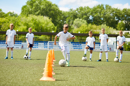 Portrait of boy in junior football team  leading ball between cones during  practice in field on sunny day with team in backgroundの写真素材