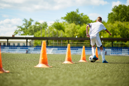 Back view portrait of teenage boy running in football field leading ball between row of orange cones during practiceの写真素材