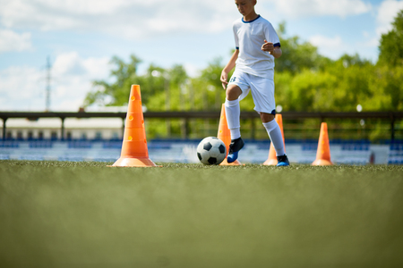 Portrait of teenage boy running in football field leading ball between row of orange cones during practiceの写真素材