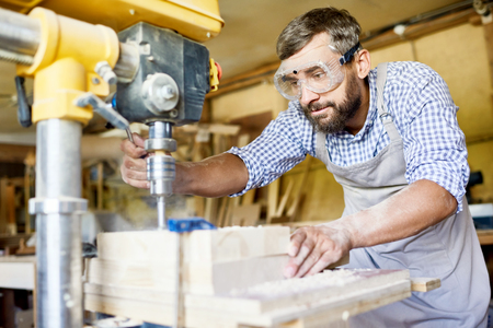 Handsome bearded carpenter wearing safety goggles and apron using drill press machine in order to make holes in wooden plank, interior of spacious workshop on backgroundの写真素材