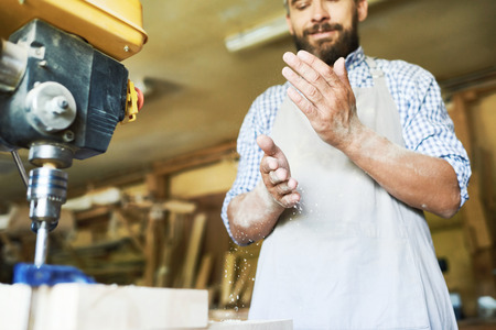 Low angle view of confident craftsman shaking sawdust from hands after completion of work with drill press machine, blurred backgroundの写真素材