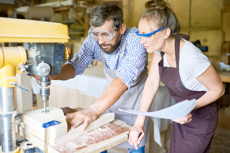 Concentrated bearded carpenter explaining his attractive apprentice how to use drill press machine, interior of spacious workshop on backgroundの写真素材