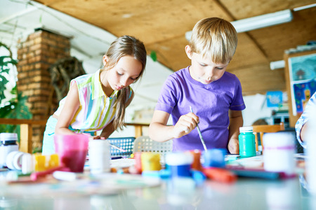 Handsome blond-haired boy and his pretty classmate wrapped up in drawing with gouache while having class at art studioの写真素材