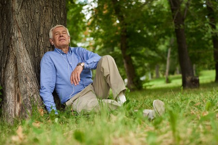Portrait of modern senior man relaxing listening to music from smartphone  in park sitting on grass with eyes closedの写真素材