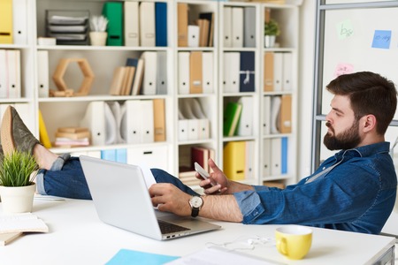 Portrait of modern bearded man in casual wear relaxing using smartphone with feet up on desk  in modern officeの写真素材