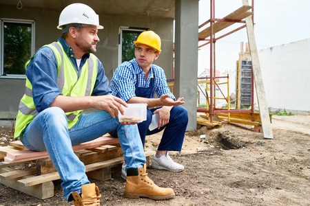 Two workers wearing protective helmets taking break from work and enjoying lunch while sitting...