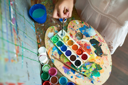 Above view image of watercolor paints tray on top of messy palette with color blots used by unrecognizable child painting on easel during art classの写真素材
