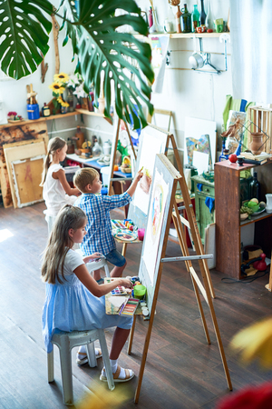 High angle portrait of three children painting on easels during art class in sunlit studio decorated with plantsの写真素材