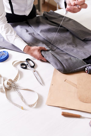 Highly professional young tailor working on custom-made male jacket with help of thread and needle...