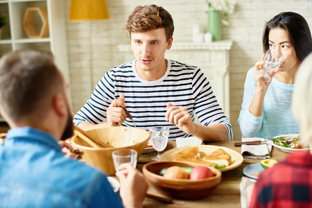 Portrait of modern young man enjoying dinner with friends sitting at big table with food and talking to peopleの写真素材