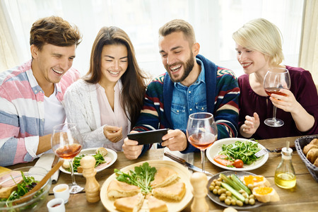 Portrait of four happy people laughing video chatting from smartphone at dinner table during holiday celebrationの写真素材
