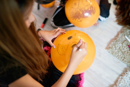 Unrecognizable little girl sitting on floor of living room and drawing scary face on orange balloon, close-up shotの写真素材