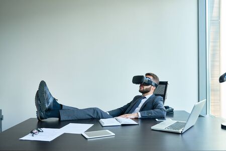 Portrait of handsome modern businessman wearing VR glasses relaxing at desk in office with feet on...