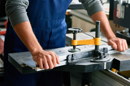 Closeup of strong male hands running piece of metal through machine unit while working in factory workshopの写真素材