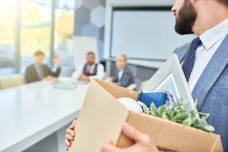 Unrecognizable man holding box of personal belongings looking back at several business people who...