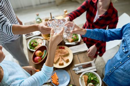 Group of friends enjoying dinner together standing at big table with delicious food and toasting, focus on clinking glasses in celebration of holidayの写真素材