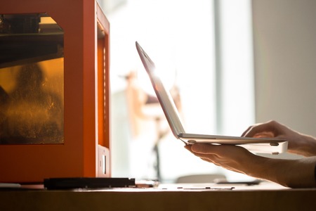 Close-up shot of unrecognizable designer sitting at office desk and working with 3D printer, lens flareの写真素材