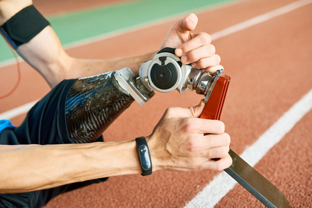 Side view  close up of unrecognizable amputee athlete fixing prosthetic leg sitting on running track in modern stadiumの写真素材